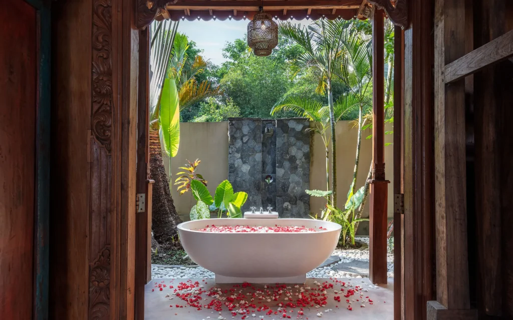 A luxurious white bathtub filled with red and white flower petals in a semi-outdoor tropical bathroom at Bali Hidden Villas.