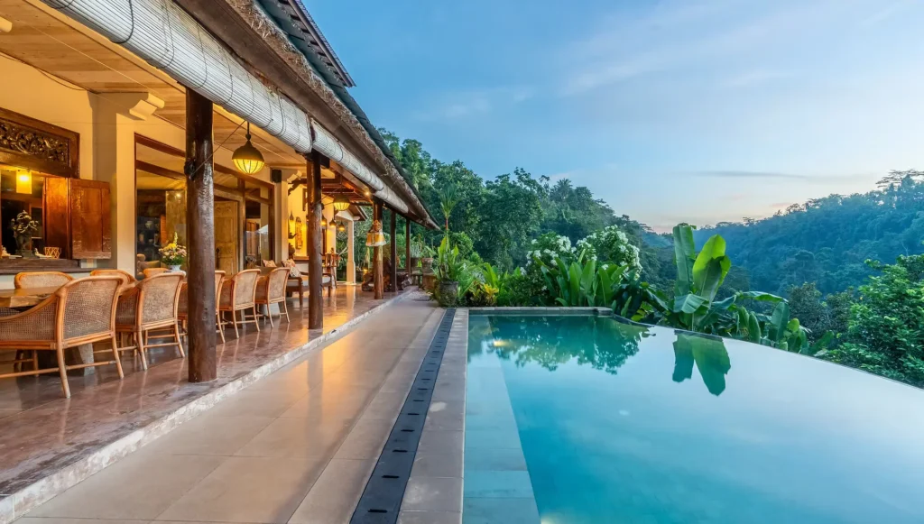 A sunrise view over a large infinity pool and traditional dining veranda overlooking a lush jungle valley at a Bali Hidden Villas property in Ubud.